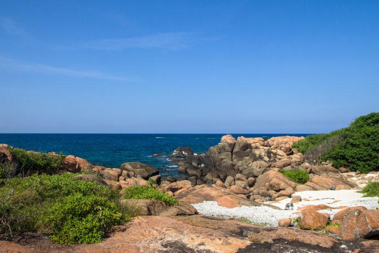 Orange granite boulders and a white coral-rubble beach on Pigeon Island, with green scrub and deep blue sea beyond