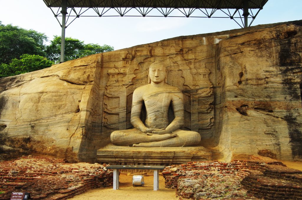 Large seated Buddha carved into a tall granite rock face at Gal Vihara, with a carved backdrop and a protective roof above