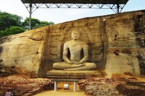 Large seated Buddha carved into a tall granite rock face at Gal Vihara, with a carved backdrop and a protective roof above