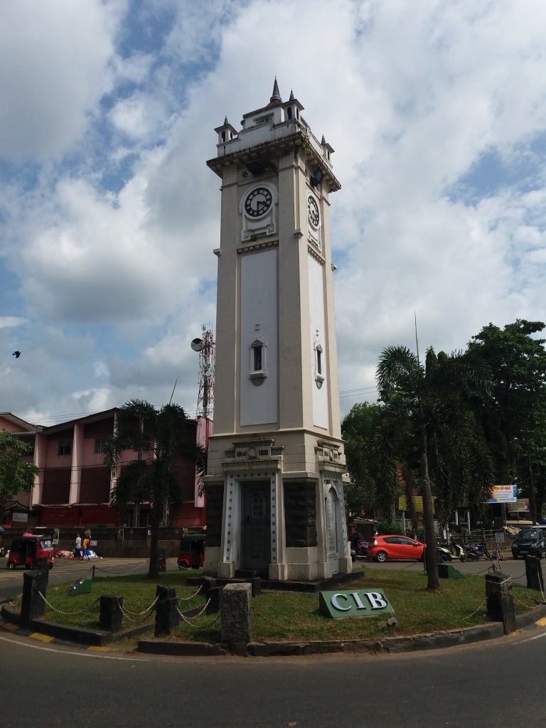 The white clock tower on a roundabout in the centre of Ratnapura, with palm trees, shops and tuk-tuks around it