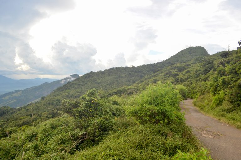 A narrow paved road running along a scrub-covered ridge towards a summit topped with transmission towers, under a cloudy sky