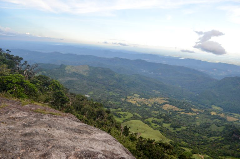 View from a bare rock on the Riverston ridge over forested slopes and a green valley with patchwork fields, with layers of hills fading into haze