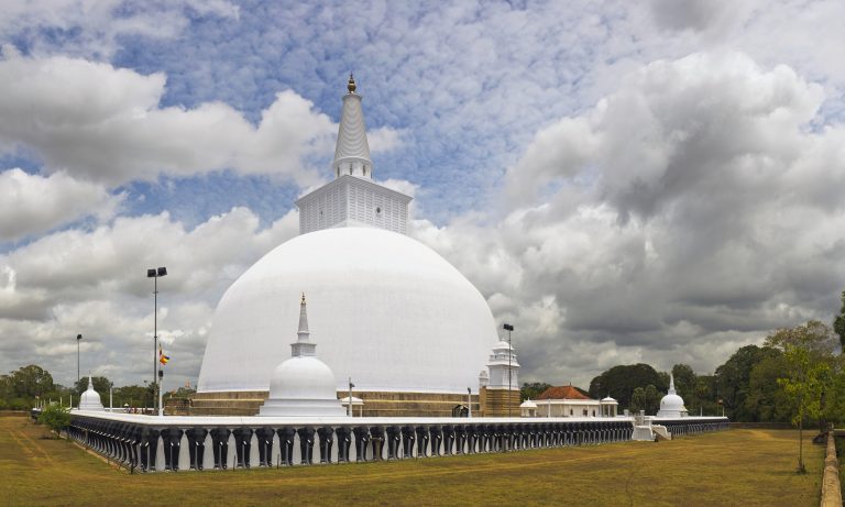 The white dome and spire of Ruwanwelisaya stupa above a wall of elephant sculptures, on a lawn under a cloudy sky