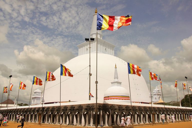 The white dome and spire of Ruwanwelisaya above a platform wall of elephant heads, with Buddhist flags flying and pilgrims in white walking by