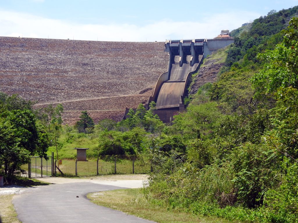 The rock-fill wall of the Samanala Dam with its concrete spillway, seen from a road through green scrub