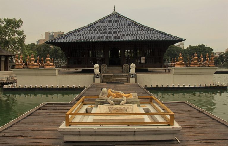 Seema Malaka pavilion with its blue-tiled roof on a platform in Beira Lake, a reclining Buddha statue on the walkway in front and rows of seated Buddhas along the platform edge