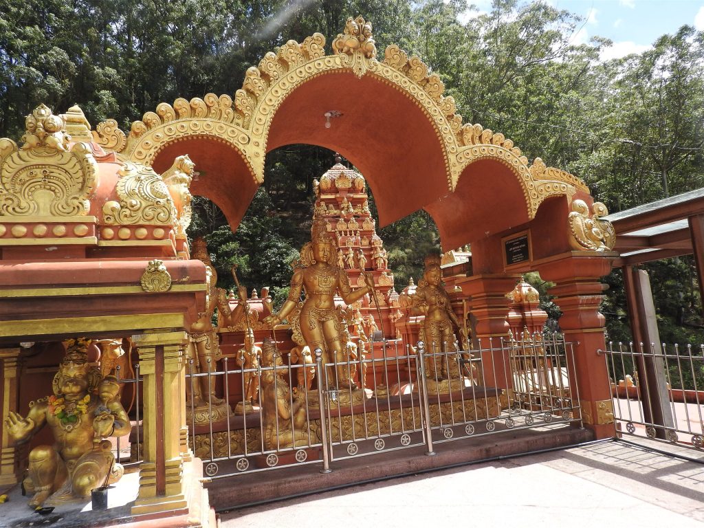 Gilded standing figures behind a railing under an ornate gold-edged arch, with a small Hanuman shrine at the left