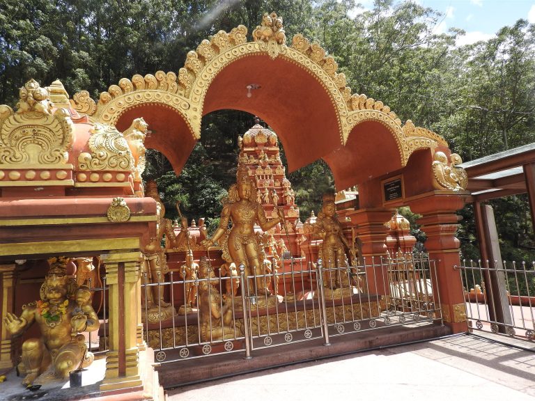 Gilded standing figures behind a railing under an ornate gold-edged arch, with a small Hanuman shrine at the left
