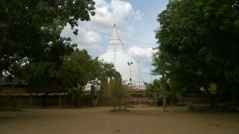 The white dome and spire of Seruwila stupa rising behind trees, with steps up to its terrace from a sandy courtyard