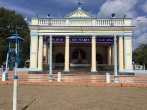 The cream-and-blue columned front of Madhu church, with 'Ave Maria' signs in Tamil, English and Sinhala, a paved forecourt and a blue bell tower on the left