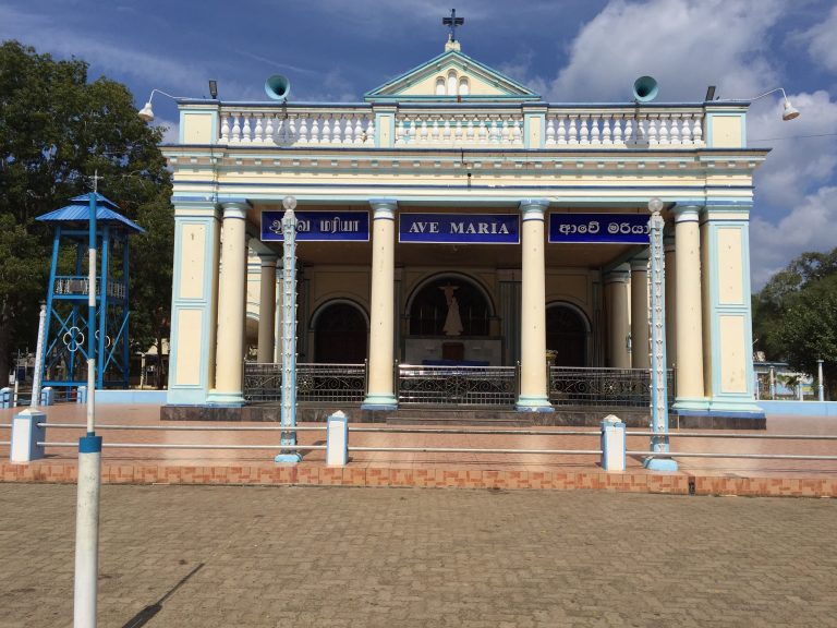 The cream-and-blue columned front of Madhu church, with 'Ave Maria' signs in Tamil, English and Sinhala, a paved forecourt and a blue bell tower on the left