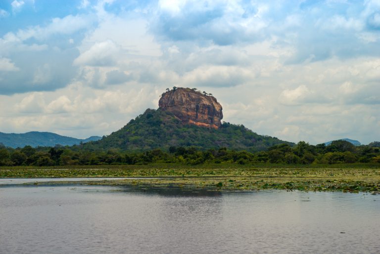 Sigiriya rock rising above forest on a green hill, seen across the lily-covered water of Thalkote Wewa under a cloudy sky