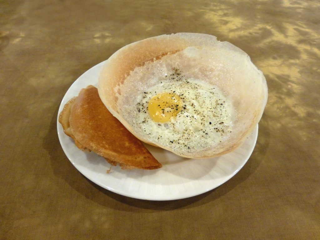An egg hopper, a thin bowl-shaped rice pancake with a fried egg in the middle, on a white plate beside a folded hopper
