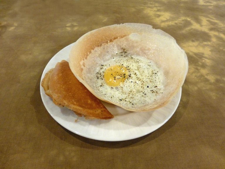 An egg hopper, a thin bowl-shaped rice pancake with a fried egg in the middle, on a white plate beside a folded hopper
