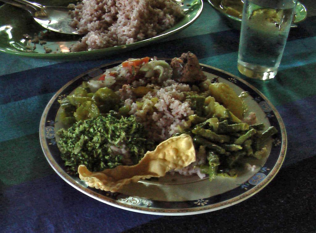 A plate of red rice with green bean curry, chopped green leaves, potato, chicken, salad and a papadum