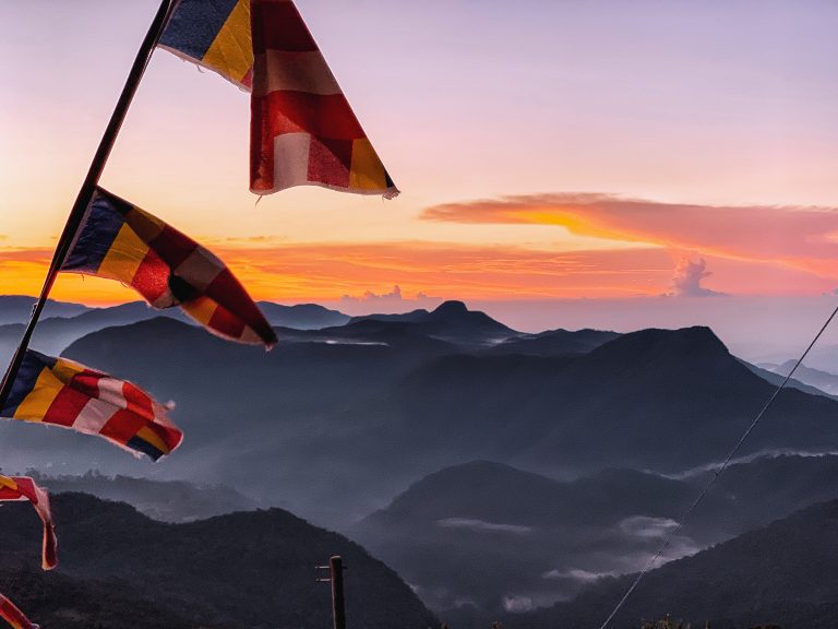 Buddhist flags in the foreground and layers of misty forested ridges under an orange sunrise sky