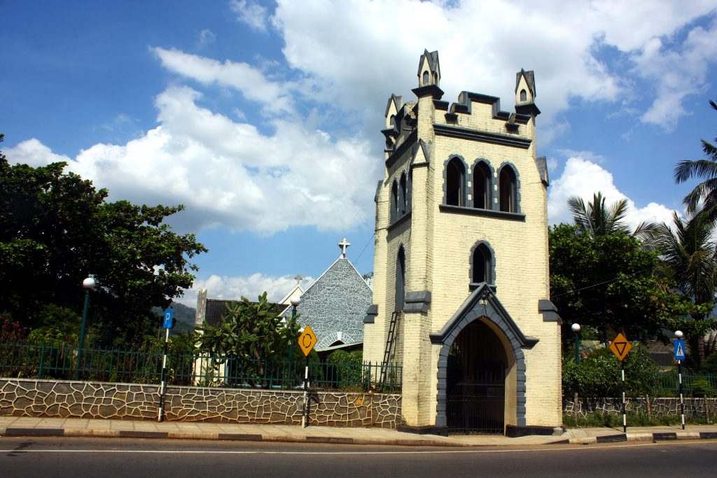 The pale square bell tower of St Mark's Church beside the road in Badulla, with the grey stone church roof and a cross behind it