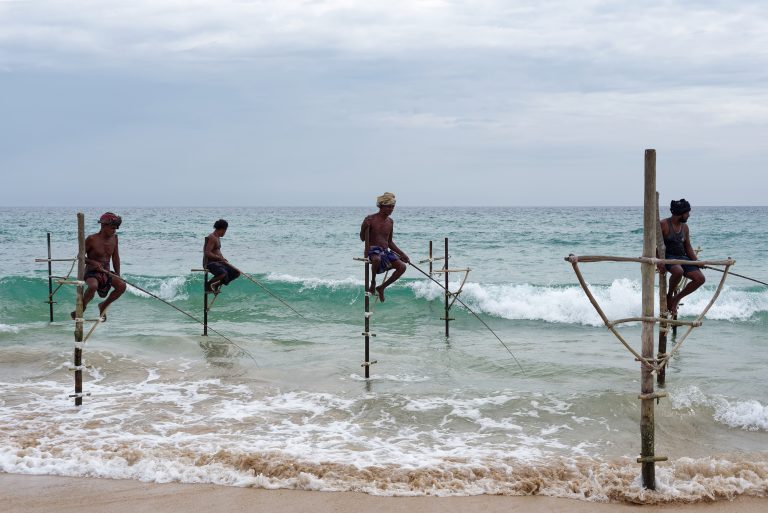 Four stilt fishermen perched on poles in the surf at Koggala, with waves breaking around them under a cloudy sky