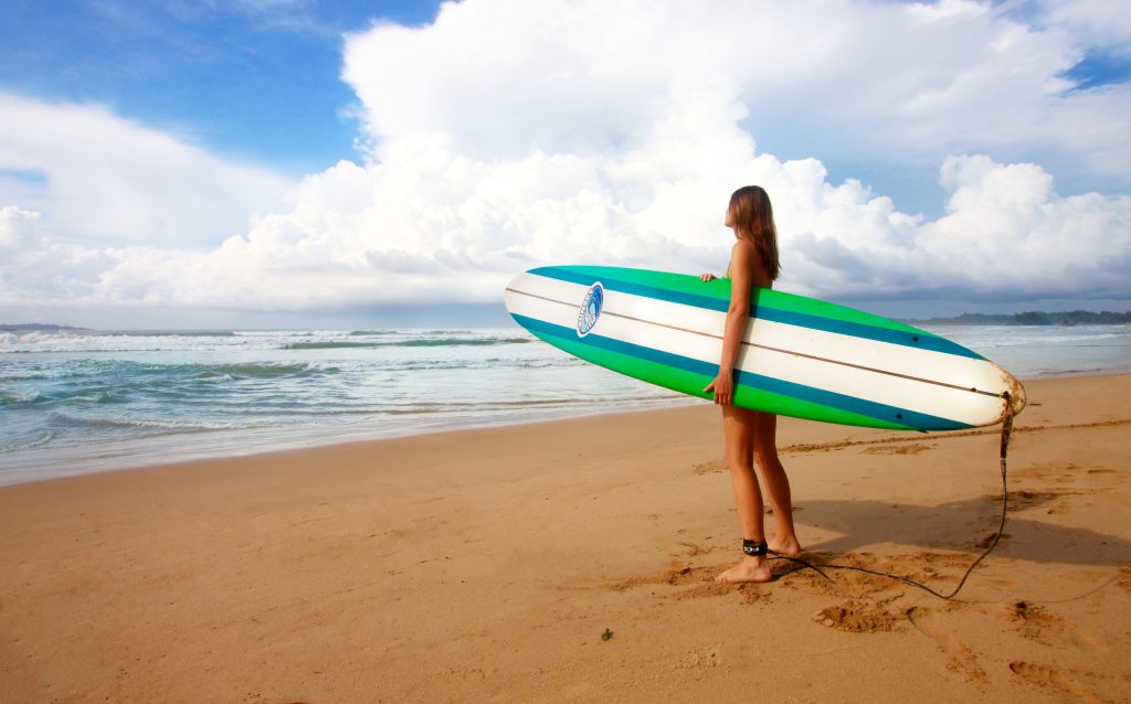 A surfer seen from behind on Weligama beach, holding a long green and white surfboard with the leash trailing on the sand, looking out at small waves under big white clouds