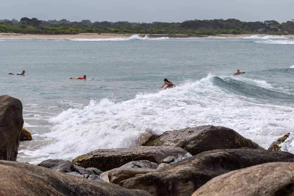 Surfers paddling and riding a breaking wave in green-blue water off Panama Beach, with dark boulders in the foreground and a low wooded shore behind