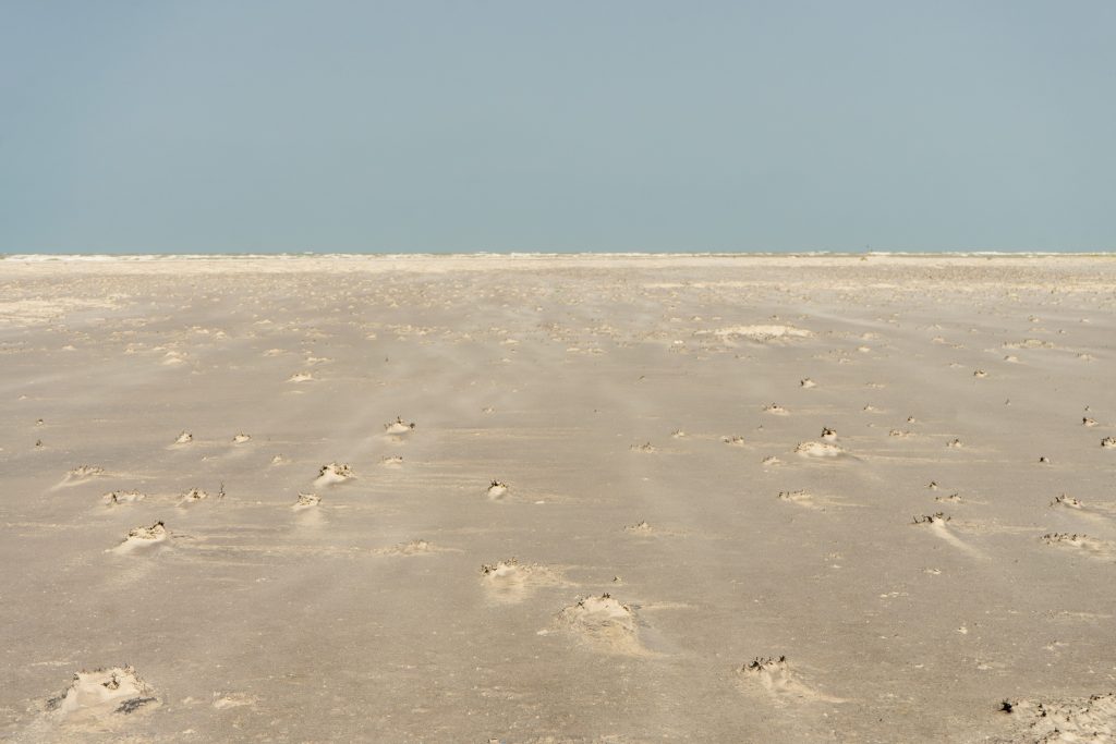 Flat, wind-rippled sand stretching to a pale sea and hazy horizon under a grey-blue sky