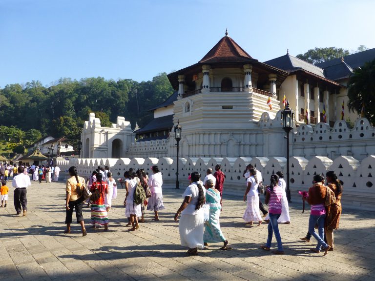 Barefoot worshippers, many dressed in white, crossing the stone forecourt in front of the octagonal tower and white wall of the Temple of the Tooth in Kandy