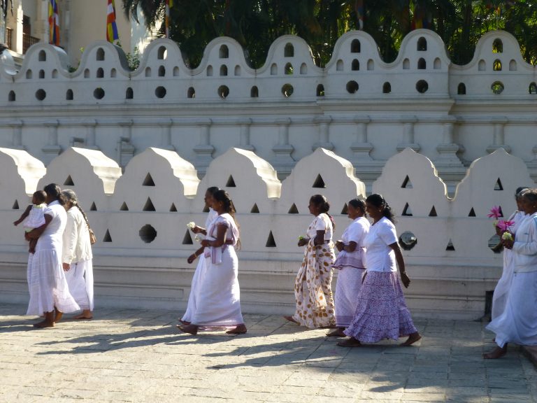 Women in white and pale saris and skirts walking barefoot beside the scalloped white wall of the Temple of the Tooth, some carrying lotus flowers