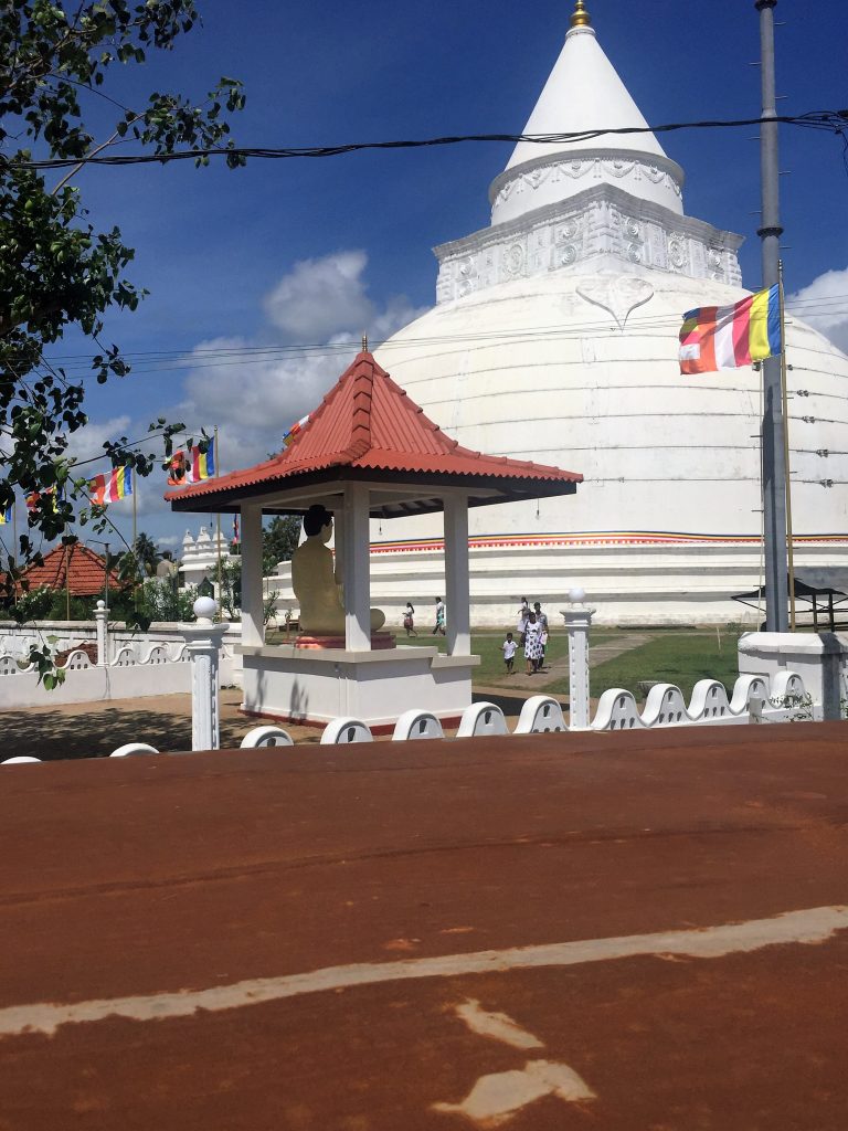 The whitewashed Tissamaharama stupa behind a small roofed Buddha shrine, with Buddhist flags and a few visitors walking on the lawn