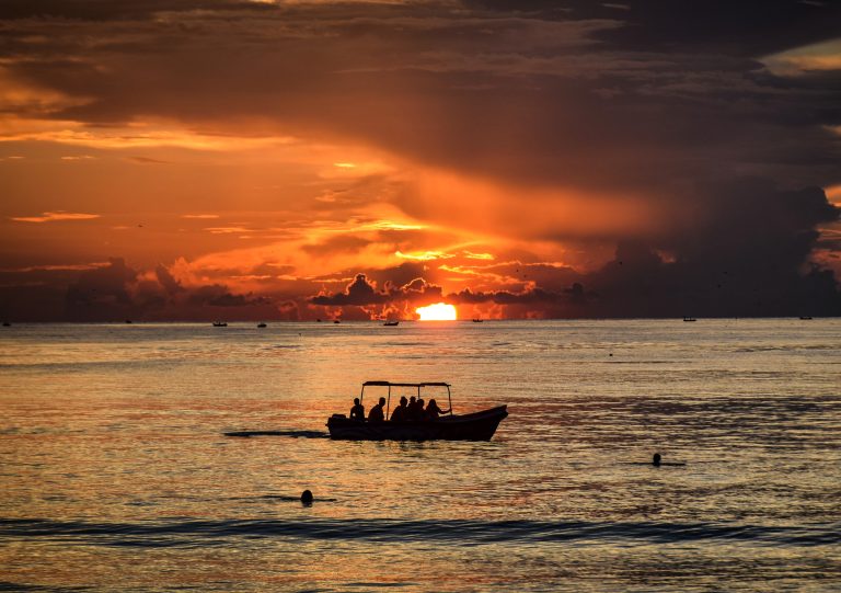 A small open boat with a canopy and a handful of passengers, silhouetted against an orange sunrise on a calm sea off Trincomalee, with fishing boats on the horizon