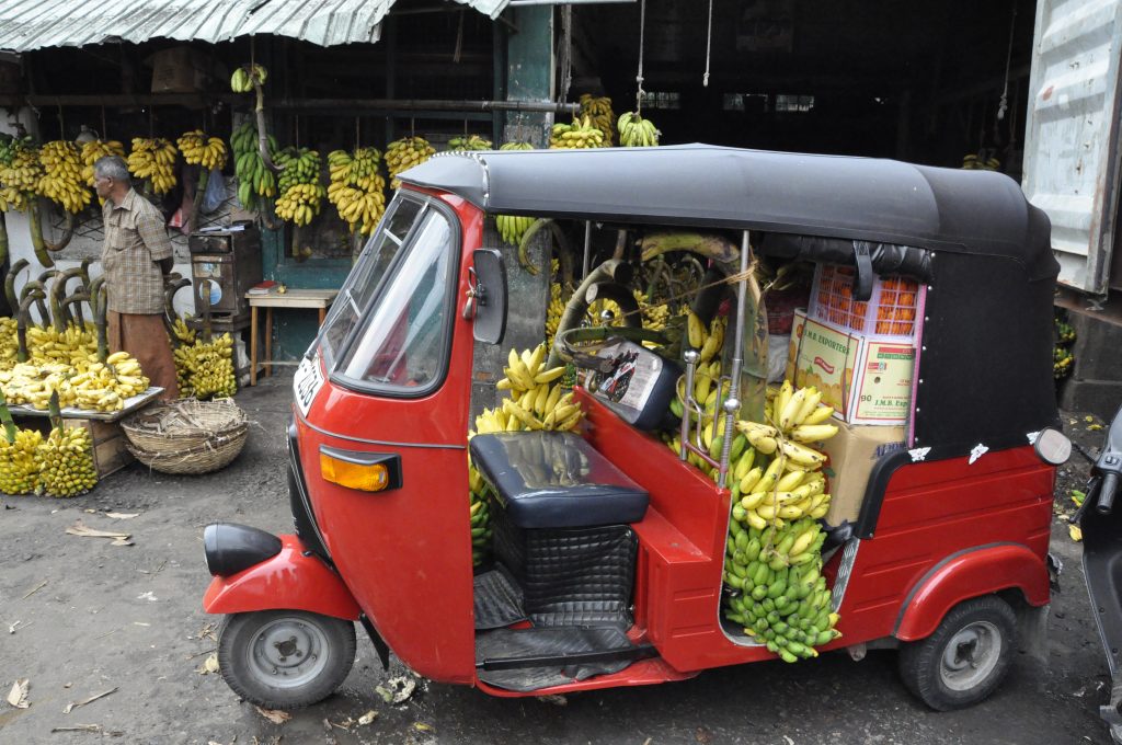 A red tuk-tuk parked at a market in Colombo, its back seat piled with bunches of bananas, with more bananas hanging at the stalls behind