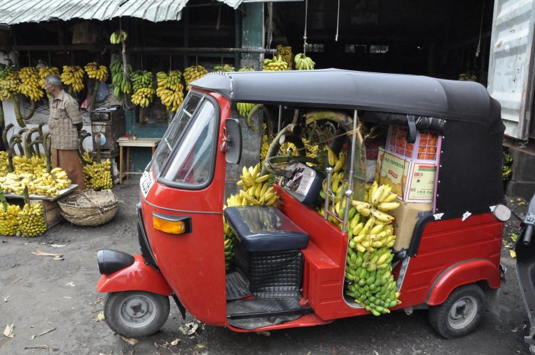 A red tuk-tuk parked at a market in Colombo, its back seat piled with bunches of bananas, with more bananas hanging at the stalls behind