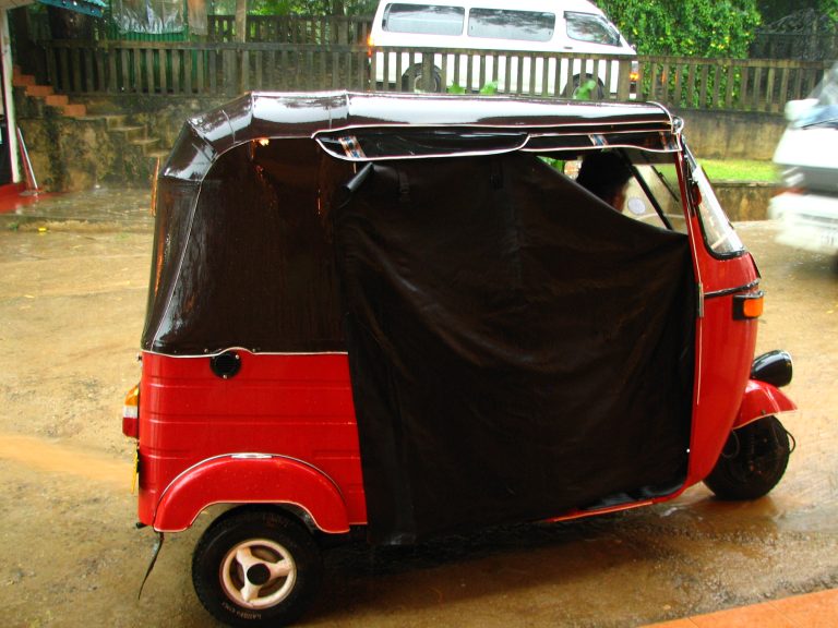 A red tuk-tuk with its black side curtains fastened shut, parked on a wet, puddled road in heavy rain