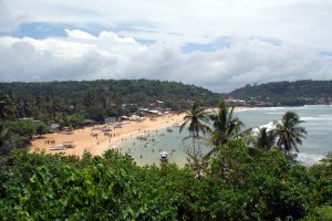 The curved sandy bay of Unawatuna seen from the hillside, with boats and swimmers in the water and wooded hills behind