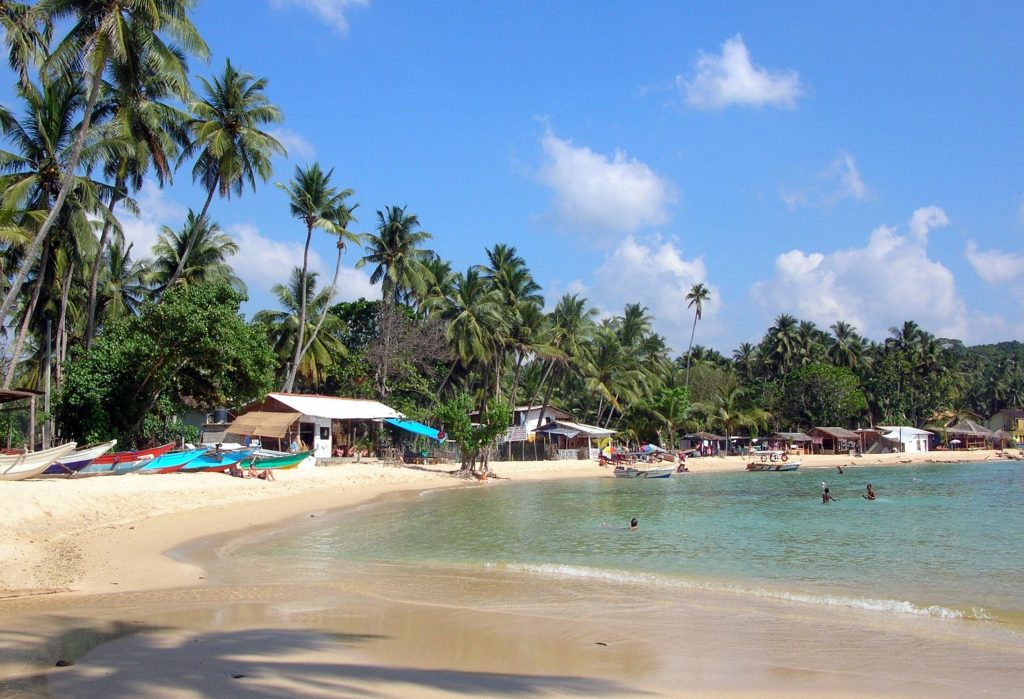 Colourful fishing boats drawn up on the pale sand at Unawatuna, with beach shacks under the palms and a few swimmers in the shallow, clear water