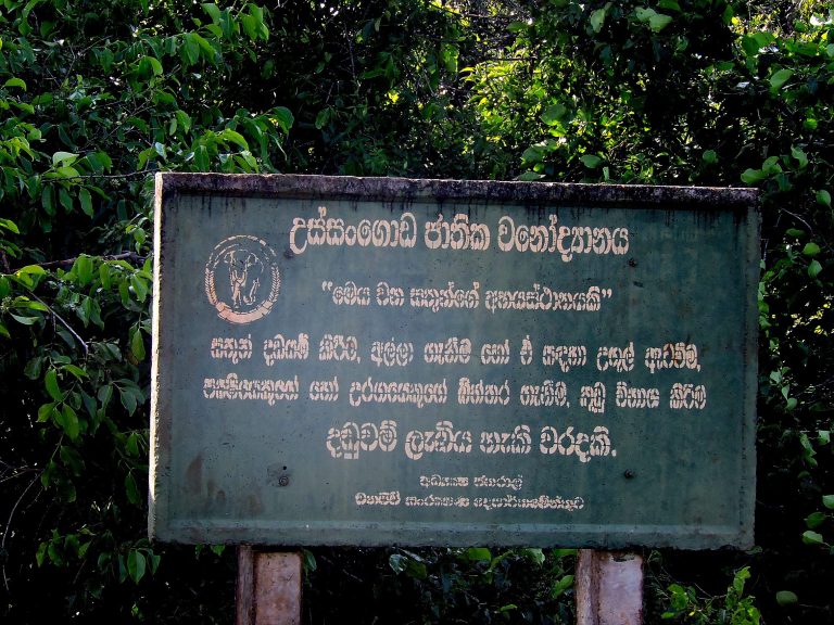 A green Department of Wildlife Conservation sign in Sinhala at the entrance to Ussangoda National Park, in front of dense foliage