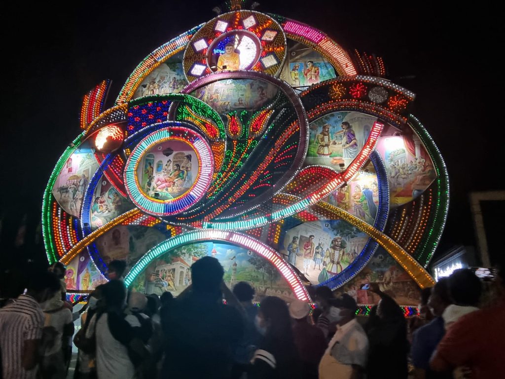A huge illuminated Vesak pandol of coloured lights and painted panels at night in Maharagama, with a crowd watching from below
