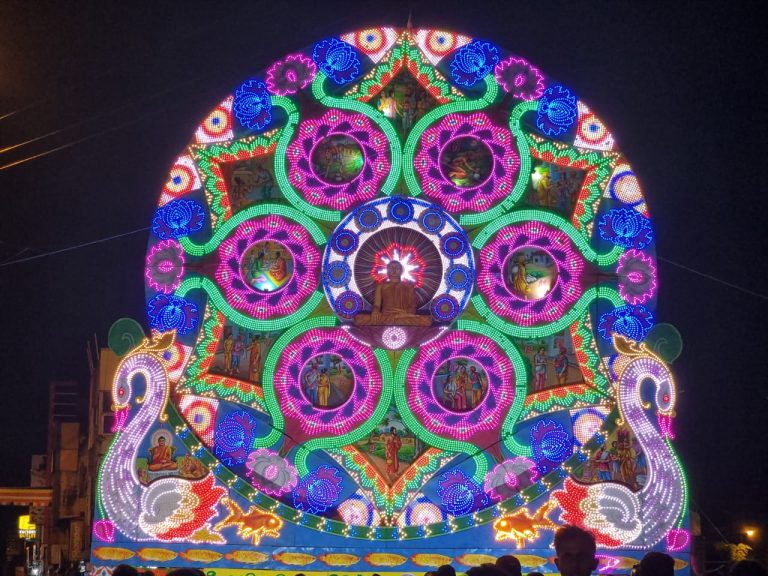 A round Vesak thorana lit with coloured lights at night in Grandpass, Colombo, with painted Buddhist scenes and swans on either side