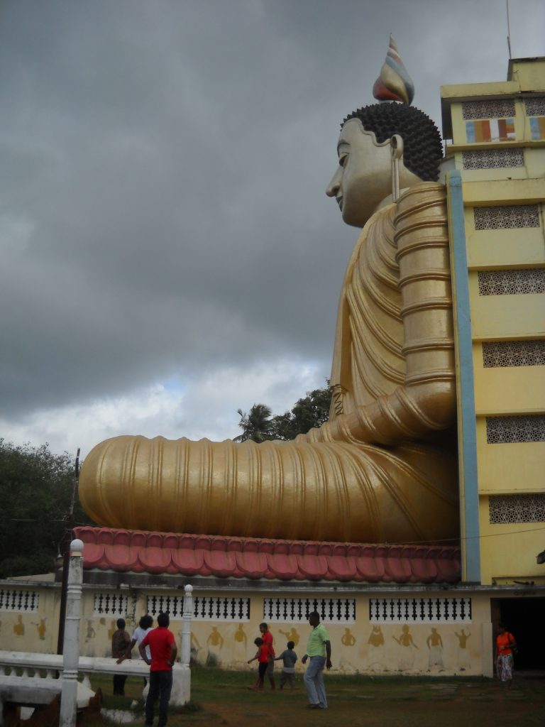 Side view of the giant seated Buddha in a gold robe against a tall yellow building, with visitors in the courtyard below