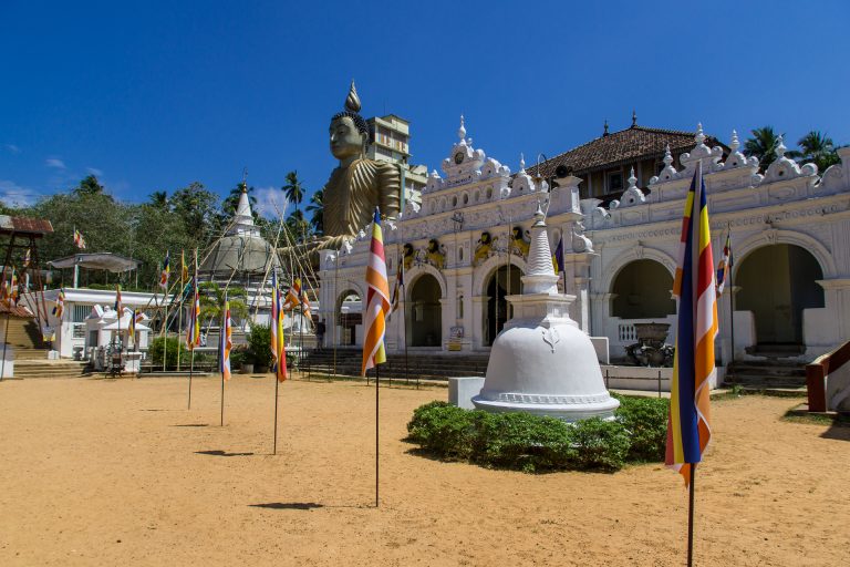 The white image house of Wewurukannala Vihara with a small stupa and Buddhist flags in a sandy courtyard, the giant seated Buddha rising behind it