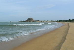 Sandy beach curving towards a large flat-topped rock outcrop on the coast, with low scrub behind